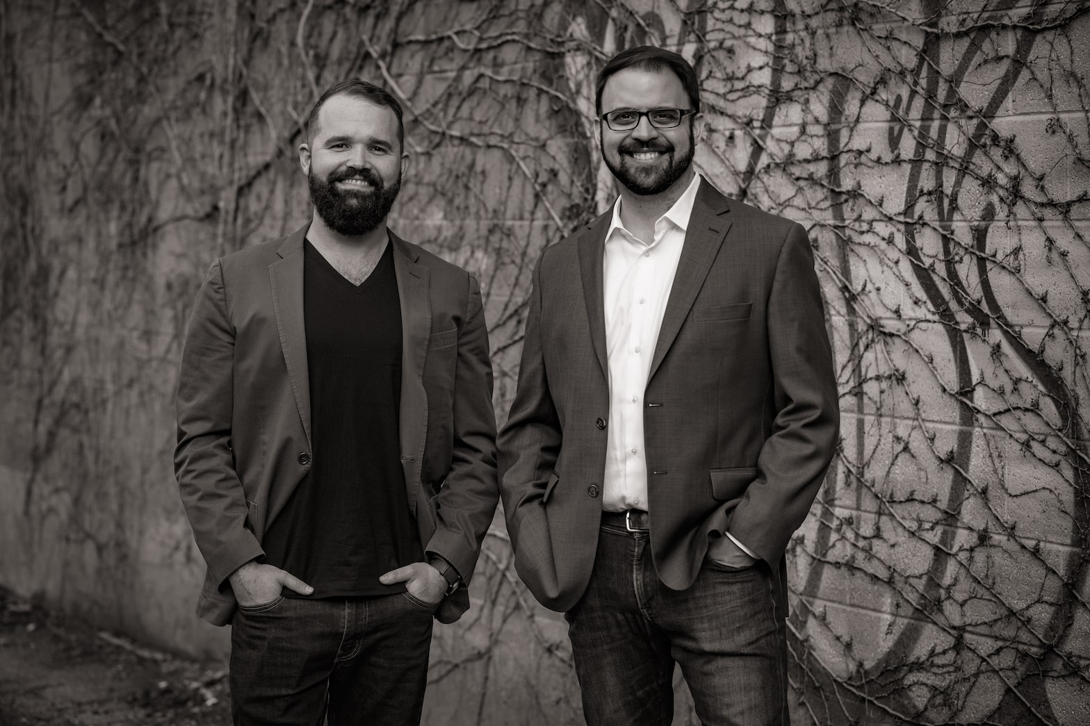 Two man smiling by vined covered brick wall.