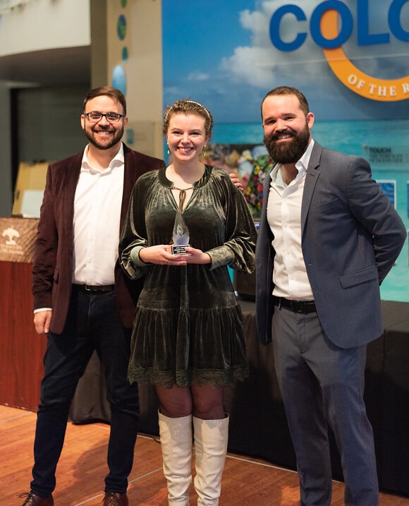 Two men and a woman stand in an aquarium in festive attire and the woman holds an award