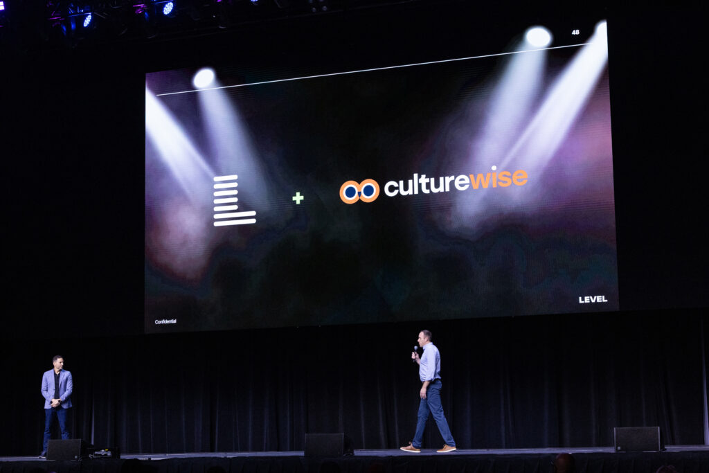 Two presenters stand on stage in front of a screen displaying the Level and CultureWise logos during a live presentation. The scene represents how Level uses CultureWise to define behaviors that strengthen its marketing agency culture for clients.