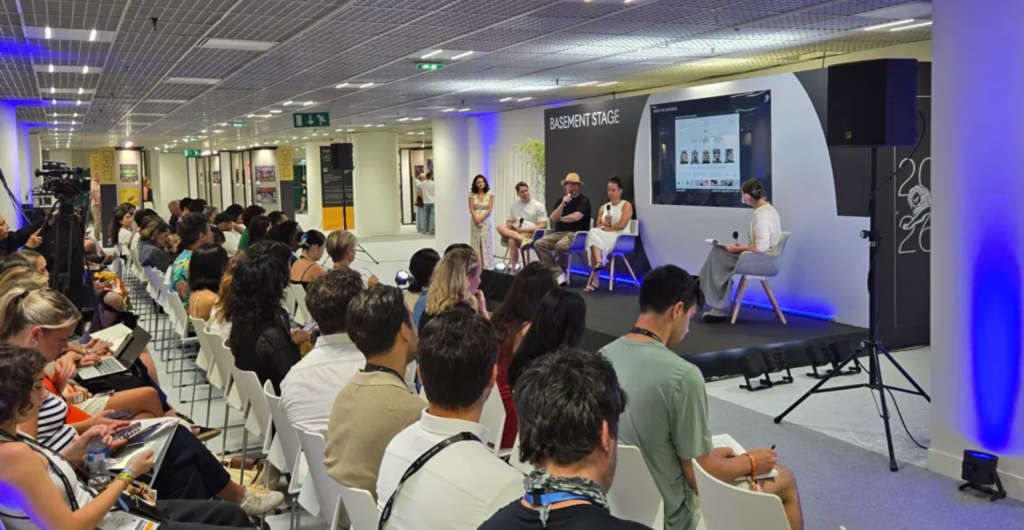 Audience seated in white chairs watching a five-person panel on the Cannes Lions Basement Stage, with a presentation screen beside the speakers.