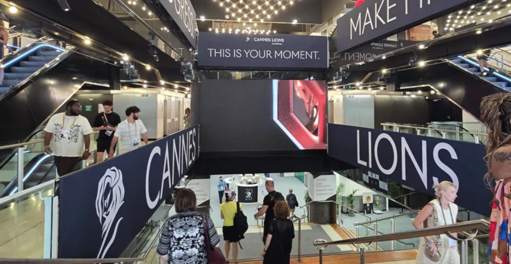 Escalators and staircase inside the Cannes Lions Palais atrium, with overhead signage reading Drive Progress, Make History, and This Is Your Moment beneath a canopy of hanging lights.