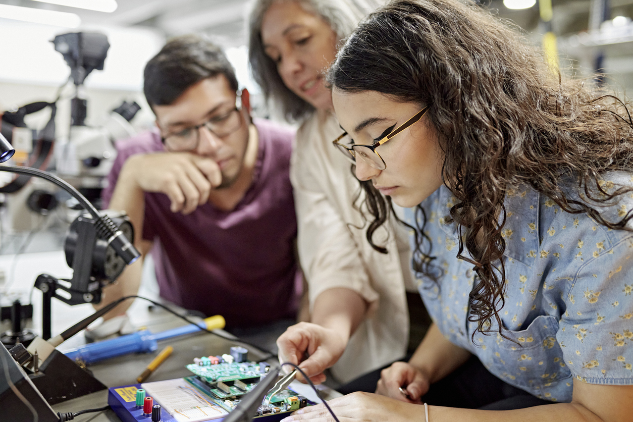 Close view of mature female educator standing between early 20s classmates and offering guidance as they work on project. Featured image for a post about the enrollment cliff.